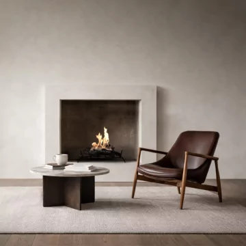 Modern minimalist living room featuring a built-in fireplace with a clean-burning flame set in a recessed concrete surround. In front, a round stone coffee table with books and a white mug sits on a textured rug beside a mid-century style brown leather lounge chair with wooden arms, creating a warm, contemporary interior setting.